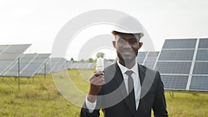 Side view of african american businessman in black suit and white helmet holding bulb while standing on solar farm