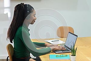 Side view at african-american female office employee using headset and laptop sitting on the workplace