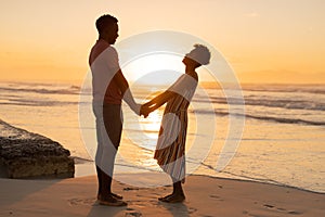Side view of african american young couple holding hands and standing on beach against sky at sunset