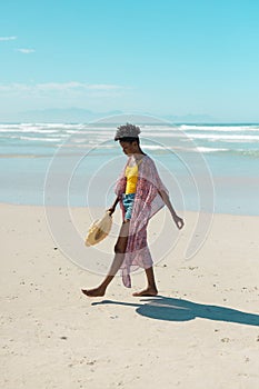 Side view of african american young woman with hat walking at beach against blue sky on sunny day