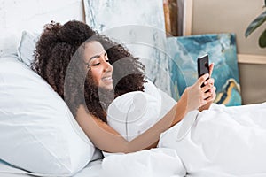 Side view of african american young woman using smartphone in bed during morning time