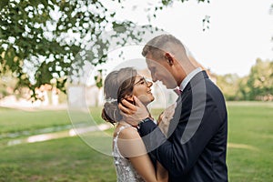 Side view of amazing wedding couple standing outside on grass in park in summer. Young groom hugging embracing bride.