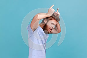 Side view of angry bully bearded man showing bull horn gesture with fingers over head.
