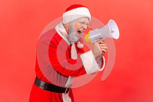 Side view of angry elderly man with gray beard wearing santa claus costume standing with megaphone