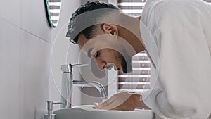 Side view arab spaniard indian arab man guy opens faucet near washbasin at home in hotel bathroom washes face with hot photo