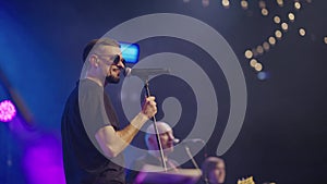 Side view artist in dark round glasses performs on club stage with band