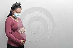 Side view of Asian pregnant woman wear face mask holding her belly, look at the stomach, isolated on grey background, COVID-19