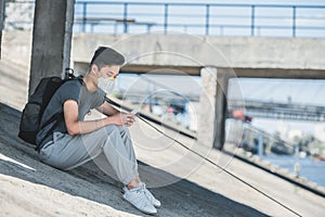 side view of asian teen in protective mask sitting under bridge and using smartphone air