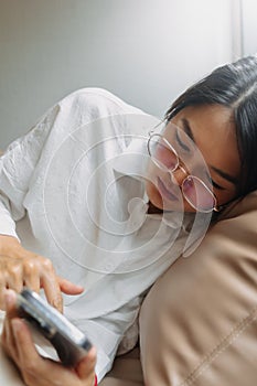 Side view of asian Thai office woman using mobile phone while leaning and resting on brown couch sofa, tired of work.