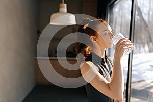 Side view of athletic young woman in sport outfit standing by window and drinking fresh water in morning after fitness