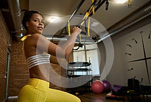 Side view of an attractive African woman doing glute exercises, performing side lunges while working out with suspension straps in