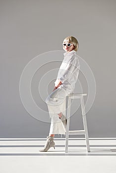 side view of attractive blonde woman in sunglasses and fashionable white outfit sitting on chair