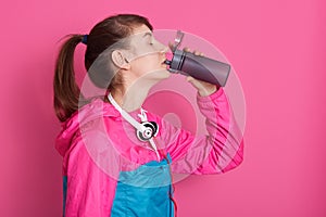 Side view of attractive brunette woman drinking water from bottle, adorable fitness girl posing isolated over pink background,