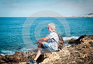 Side view of an attractive senior man casual dressed sitting in front to the sea. White hair and beard. Sunrise. Blue ocean and