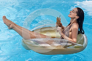 Side view of attractive slim tanned woman sitting on swimming ring, holding cocktail in hand and relaxing at summer luxury resort