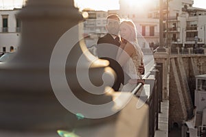 Side view of attractive wedding couple standing at steel railing of bridge at sunset. Young bride embracing with groom.