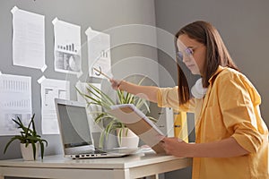 Side view of attractive woman with brown hair wearing yellow shirt posing in office, holding papers, pointing at graphs on the