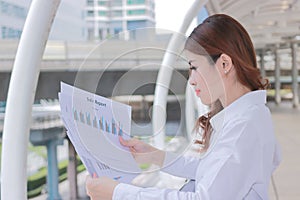 Side view of attractive young Asian business woman analyzing paperwork or charts in her hands at outside office.