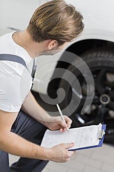 Side view of automobile mechanic writing on clipboard while examining car's wheel in workshop