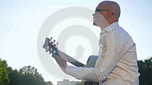 Side view of a bald man plays guitar and sings in park. Guitarist touching guitar strings. Medium shot.