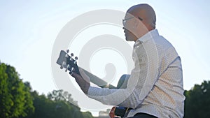 Side view of a bald man plays guitar and sings in park. Guitarist touching guitar strings. Medium shot.