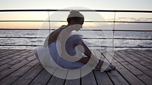 Side view of a ballerina sitting on a wooden flour outdoors near the sea or ocean. Young ballet dancer in white tutu