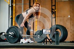 Side view, with barbell. Smartphone on tripod capturing video. Beautiful strong woman is in the gym