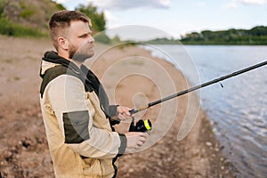 Side view of bearded fisherman holding casting rod wearing raincoat standing on bank waiting for bites on water river at