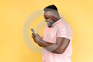 Side view of bearded happy man using mobile phone with happy expression, addicted to smartphone.