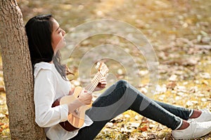 Side View of beautiful Asian woman looks away and enjoy spending leisure time playing ukulele in the park, concept musical