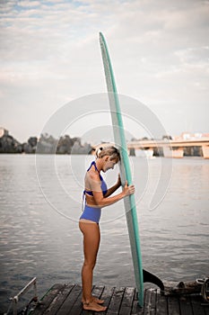 Side view of beautiful blonde woman who stands on wooden pier with surfboard in her hands