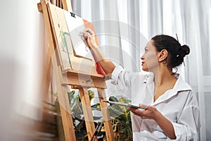 Side view of beautiful brunette painter with gathered hair in a bun and paint brushes in her hair drawing using pencil in front of