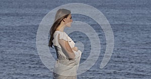 Side view of a beautiful Caucasian woman in light white dress standing on the background of sea waves and thinking