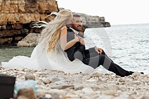 Side view of beautiful couple sitting on stones pebbles at shore near water. Young woman bride hugging kissing groom.