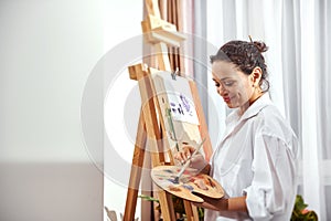 Side view of a beautiful female painter mixing oil paint on a palette and standing in front of an easel