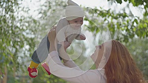 Side view of beautiful red-haired woman playing with her kid in the park or garden close-up. The lady enjoying the sunny