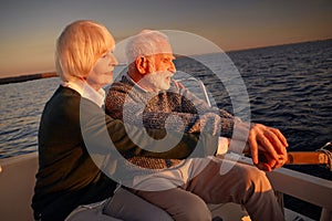 Side view of beautiful senior couple sailing at sunset, elderly man and woman sitting on the side of boat or yacht deck