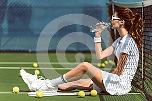 side view of beautiful tennis player in white tennis uniform and sunglasses drinking water while resting on court with racket