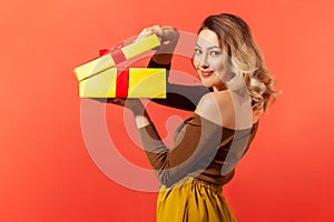 Side view beautiful woman unpacking big yellow gift box and looking at camera with pleasant smile, satisfied with present