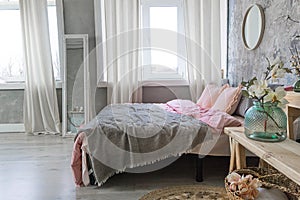 Side view on a bedroom  bed with grey and pink bedding, white mirror and vases with flowers