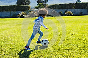 Side view of biracial boy with afro hair running and kicking soccer ball on grassy field in yard