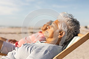 Side view of biracial senior woman sleeping while relaxing on chair with man at beach