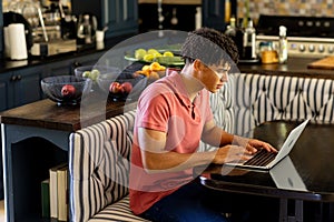 Side view of biracial young man with dreadlocks working over laptop on table at home, copy space
