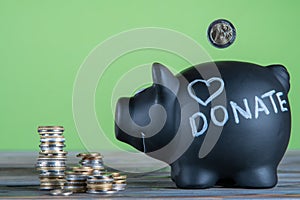 Side view of black piggy bank with word Donate and sign heart near stacks of coins on grey table on green background.