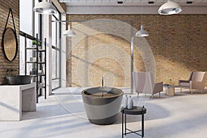 Side view on black stylish bath in sunlit spacious loft style bathroom with brick wall, armchairs on light concrete floor and