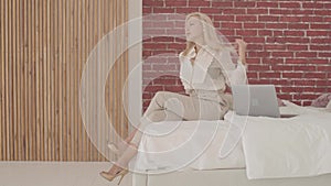 Side view of a blond Caucasian businesswoman sitting with laptop on the bed and looking away. Young beautiful lady in