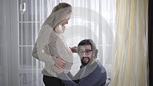 Side view of blond Caucasian woman and her husband indoors. Extremely happy man listening to his future baby`s heartbeat