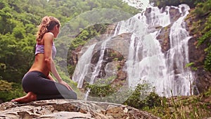 Side view blond girl sits in yoga pose in front of waterfall