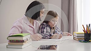 Side view of brunette Caucasian mother helping daughter to write down. Pretty schoolgirl doing homework with mom at home