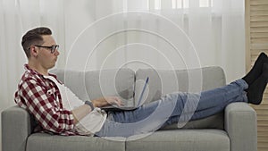 Side view of brunette man lying on couch and using laptop. Wide shot of confident Caucasian guy surfing Internet or